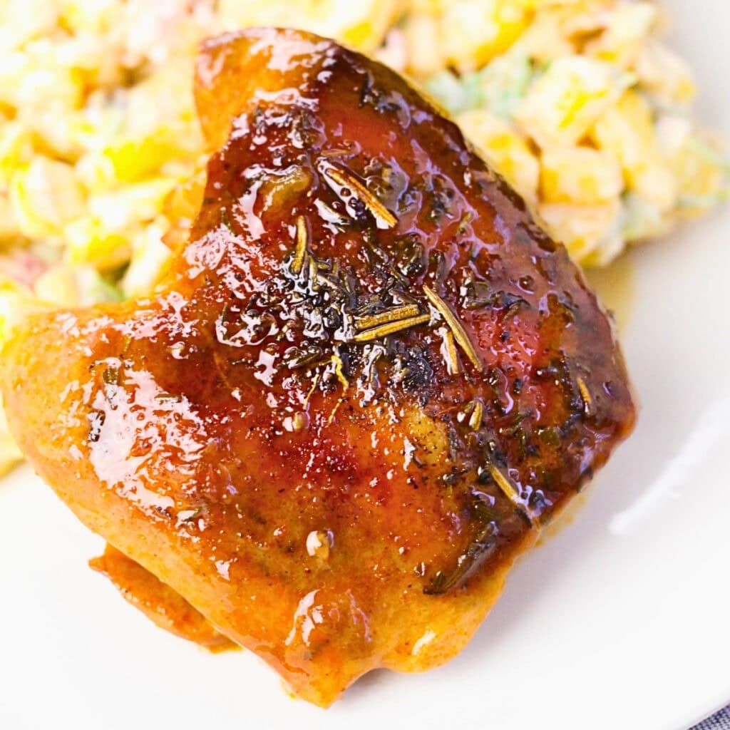 Glazed chicken thigh with herbs, served next to a creamy salad on a white plate.