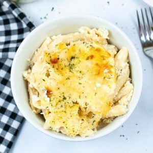 Bowl of baked cheesy pasta with herbs, next to a fork and checkered napkin.