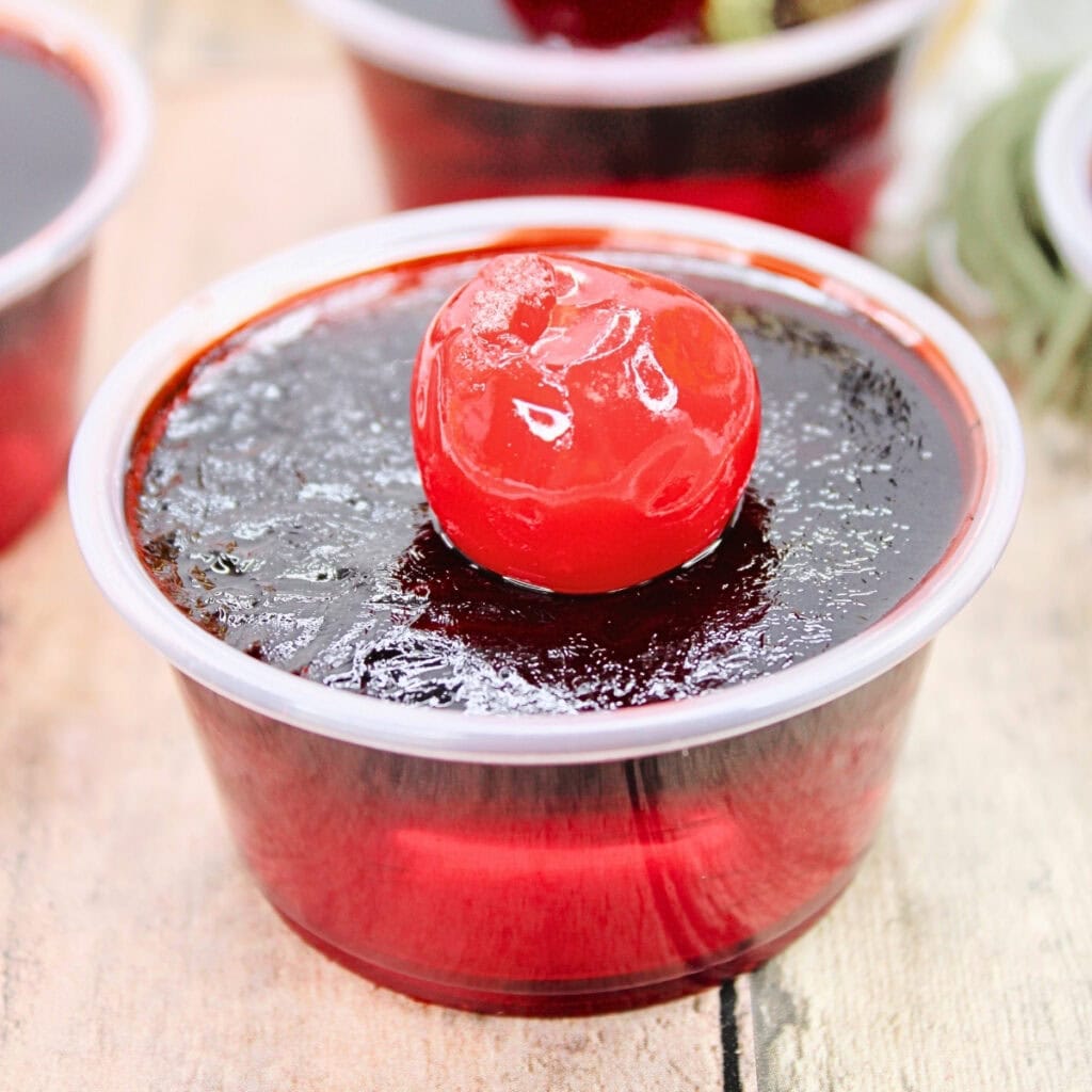 Red gelatin cup topped with a glossy maraschino cherry on a wooden surface.