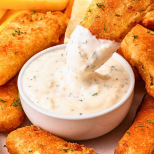Golden fried fish stick being dipped into creamy tartar sauce in a white bowl.