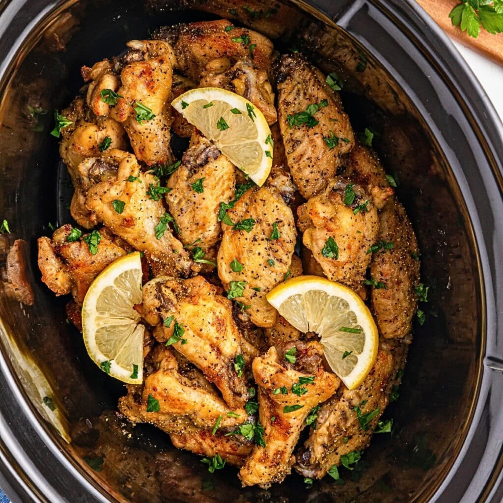 Chicken wings with lemon slices and parsley in a black slow cooker.