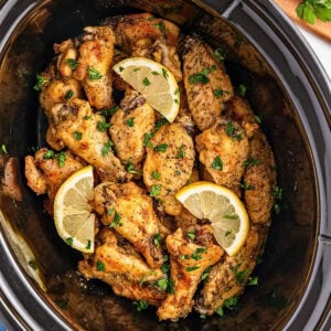 Chicken wings with lemon slices and parsley in a black slow cooker.
