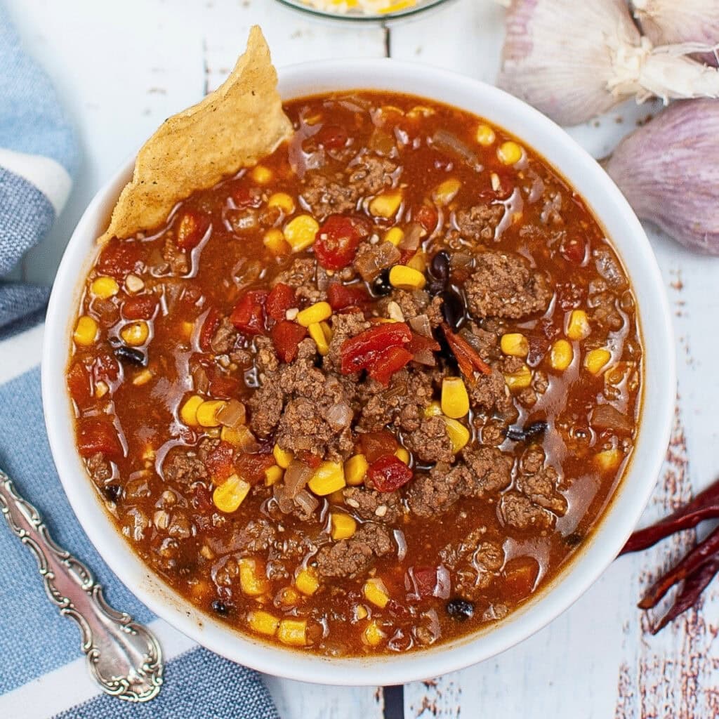 Bowl of chili with corn, ground beef, tortilla chip, and visible seasonings.