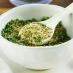 White bowl of mixed dried herbs with a white spoon scooping some out.