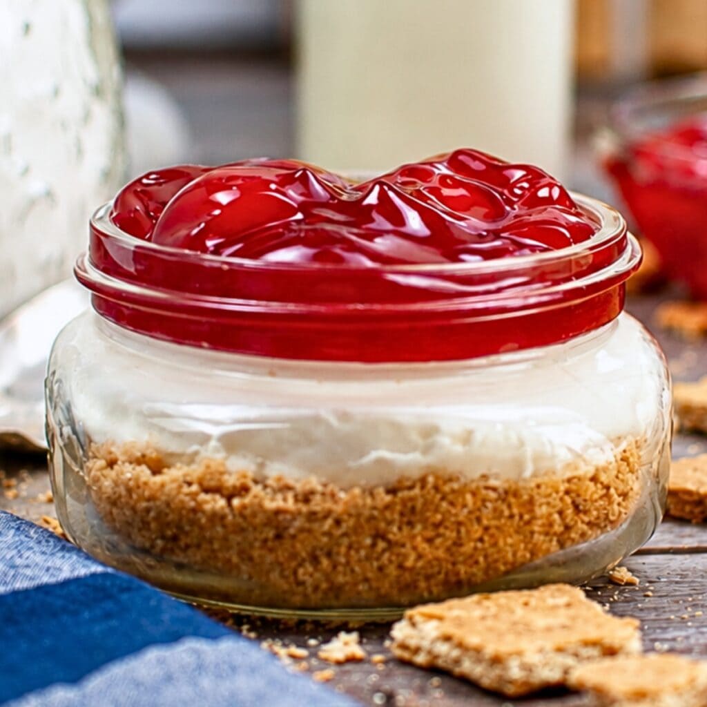 Mini cheesecake in a jar topped with cherry pie filling and graham cracker crust.