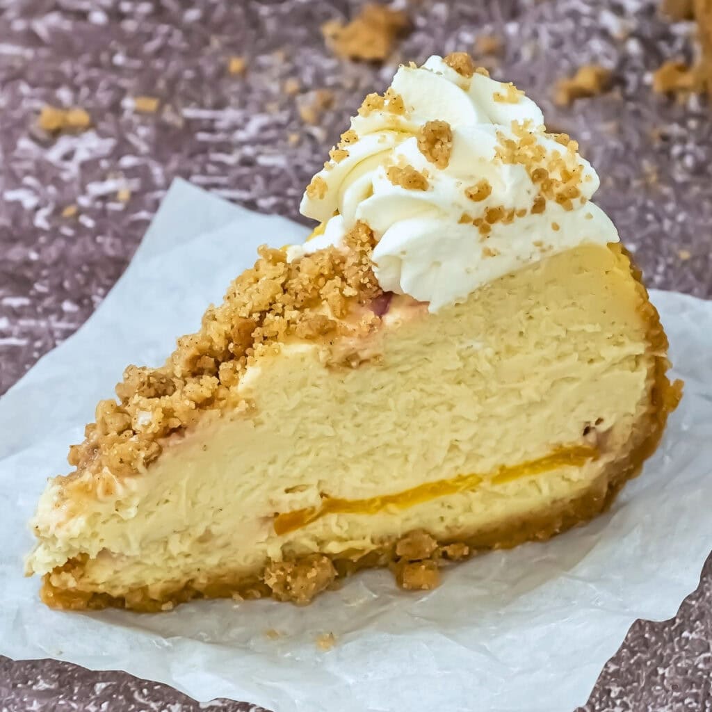 Slice of cheesecake with whipped cream and crumb topping on parchment paper.