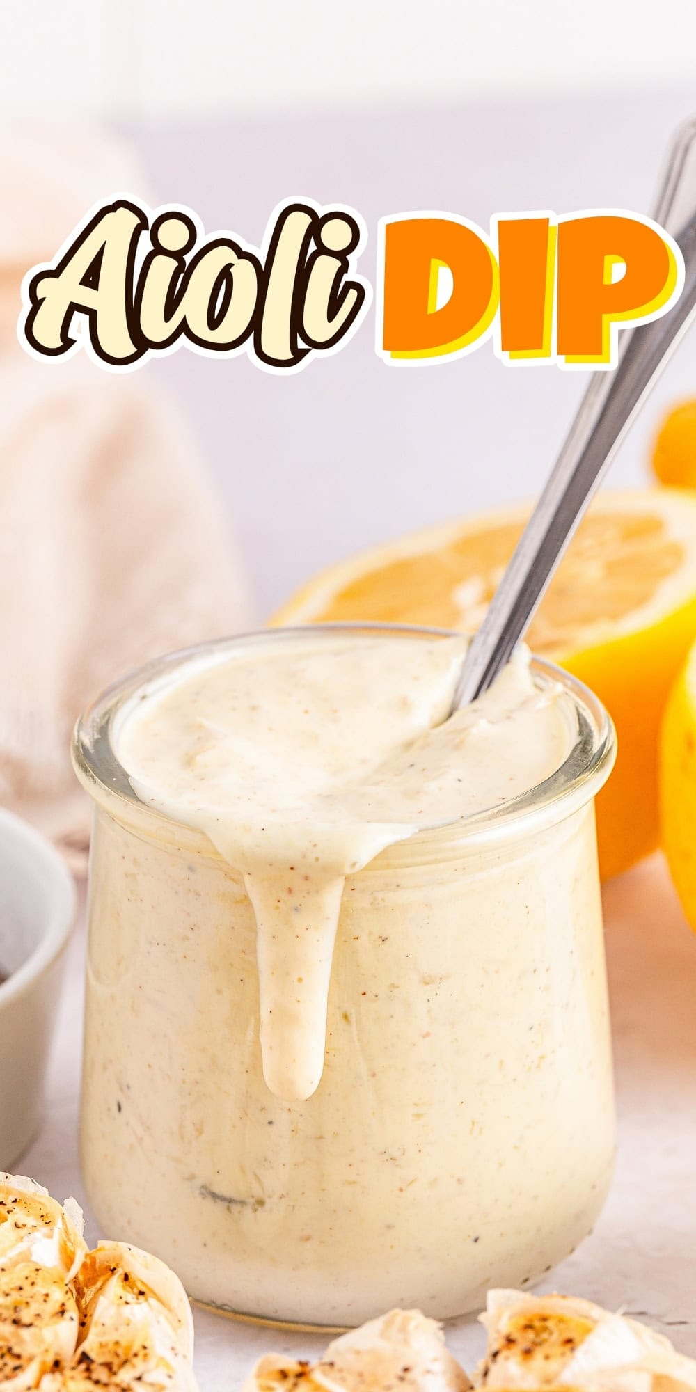 Glass jar of creamy aioli dip with spoon, lemons, and garlic beside it.