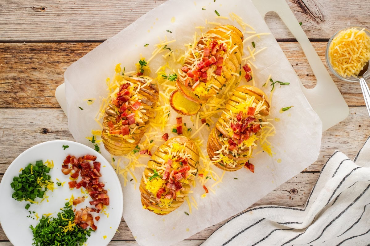 Sliced baked potatoes topped with cheese, bacon, and herbs on parchment.