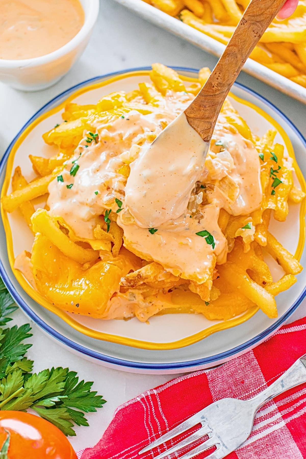 French fries topped with creamy sauce, parsley, and a wooden spoon on a plate.