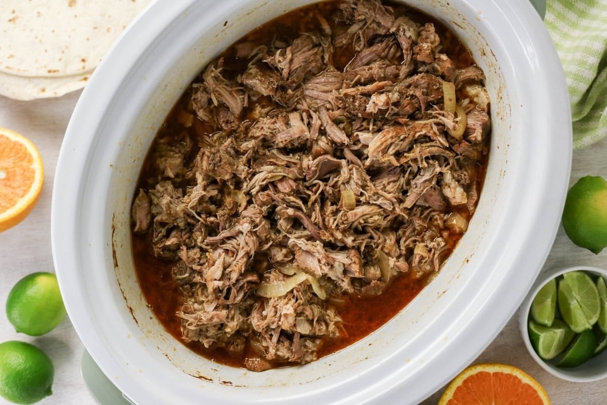 Shredded beef in sauce inside a slow cooker, surrounded by limes and oranges.