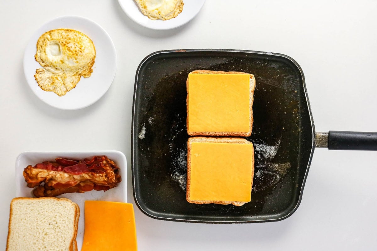 Two sandwiches with cheese cooking in a pan, with eggs and bacon nearby.