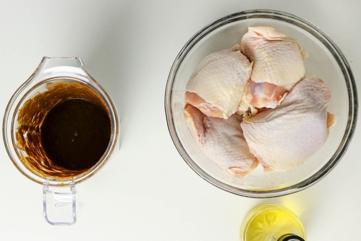 Raw chicken thighs in a bowl next to a bowl of marinade and a bottle of oil.