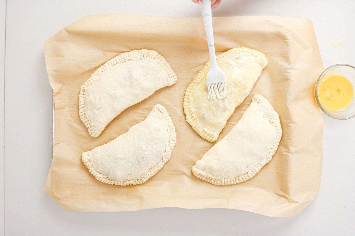 Hand brushing egg wash on uncooked empanadas on parchment-lined baking tray.