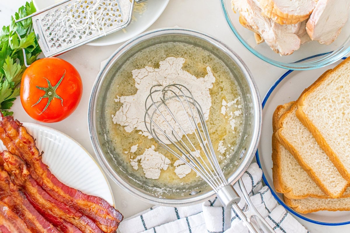 Whisk mixing flour and butter in bowl, surrounded by bread, bacon, tomato.