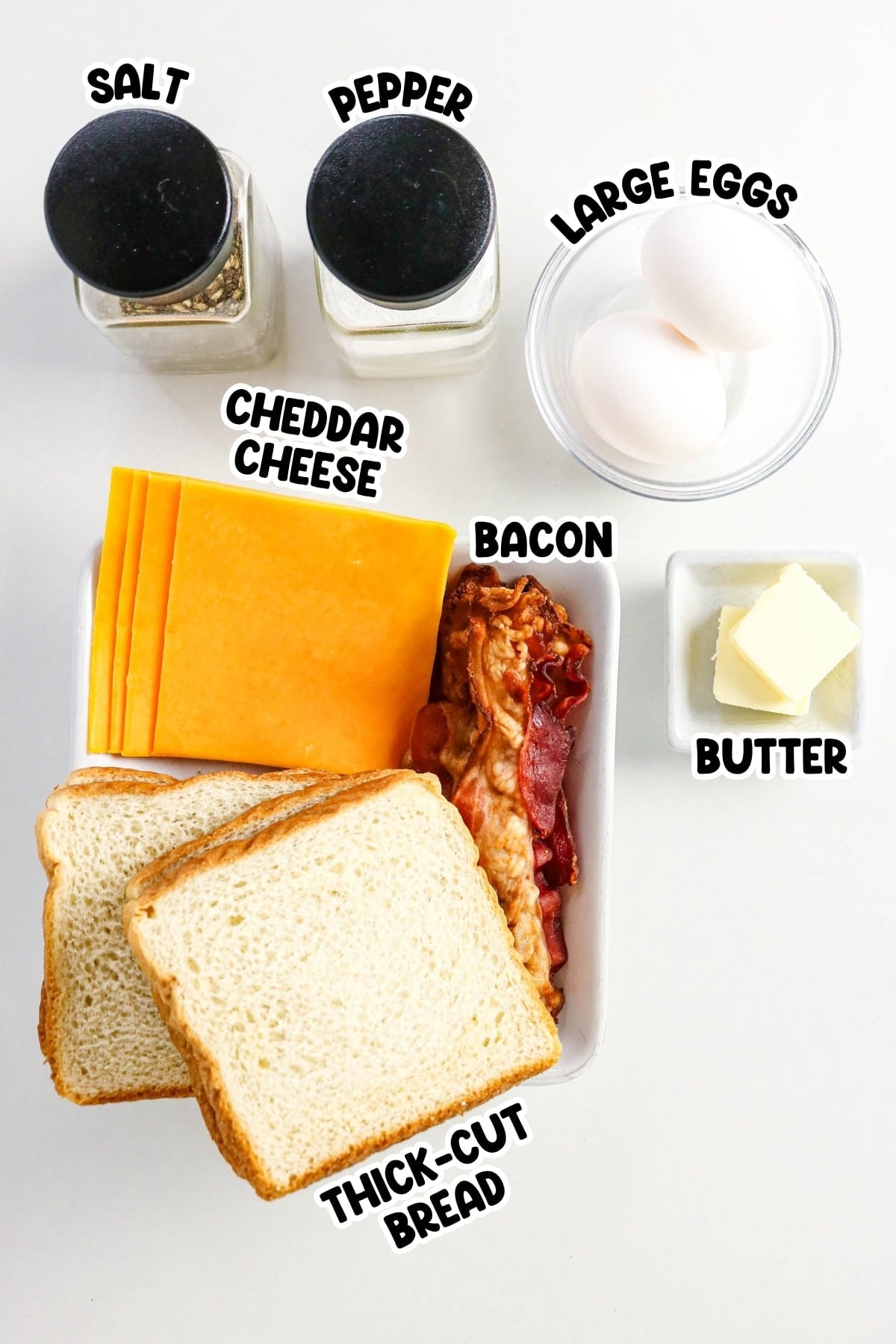 Bread, cheddar, bacon, eggs, butter, salt, and pepper on a white surface.