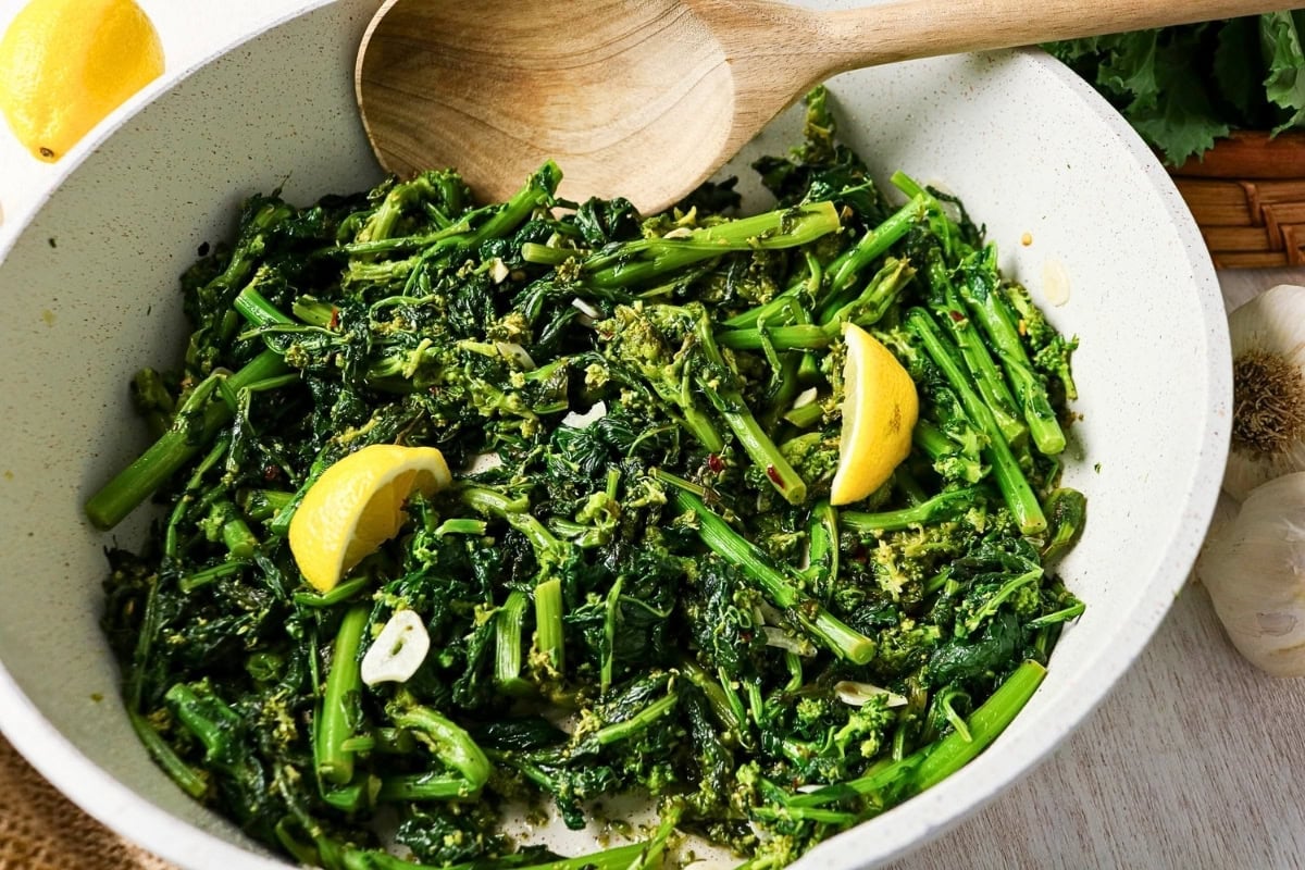 Saut&eacute;ed broccoli rabe with lemon wedges in a white pan with a wooden spoon.