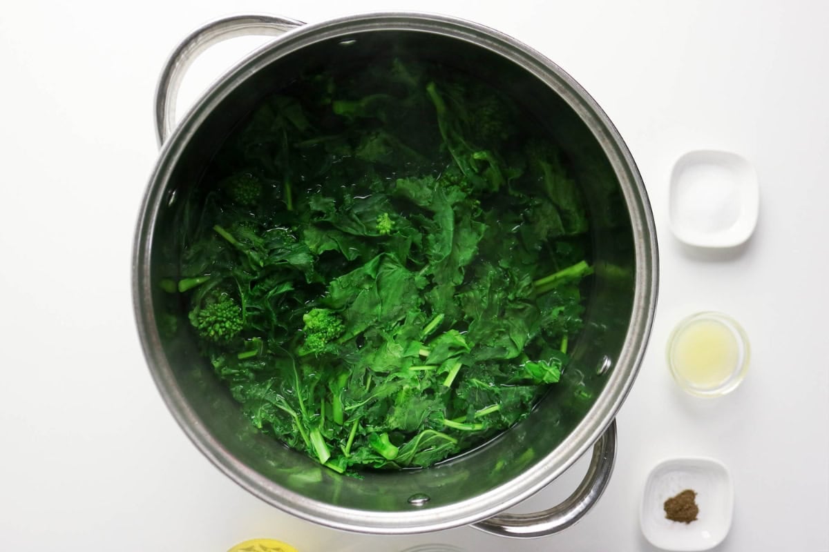 Green vegetables boiling in a large pot, with seasonings in small bowls nearby.