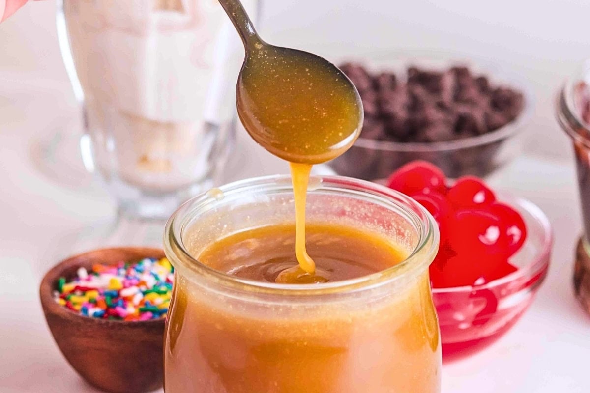 Spoon drizzling caramel sauce into a jar, with sprinkles and cherries nearby.