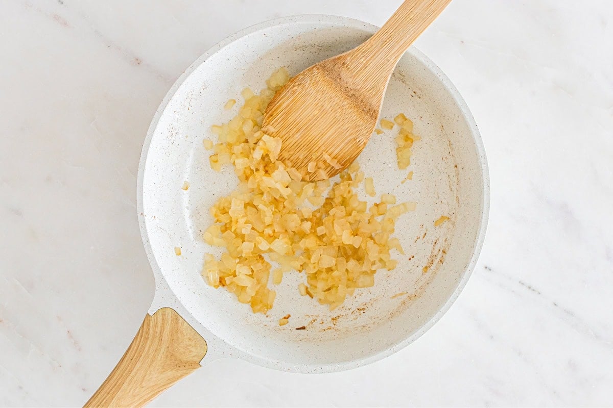 Diced onions being sautéed in a white pan with a wooden spatula.