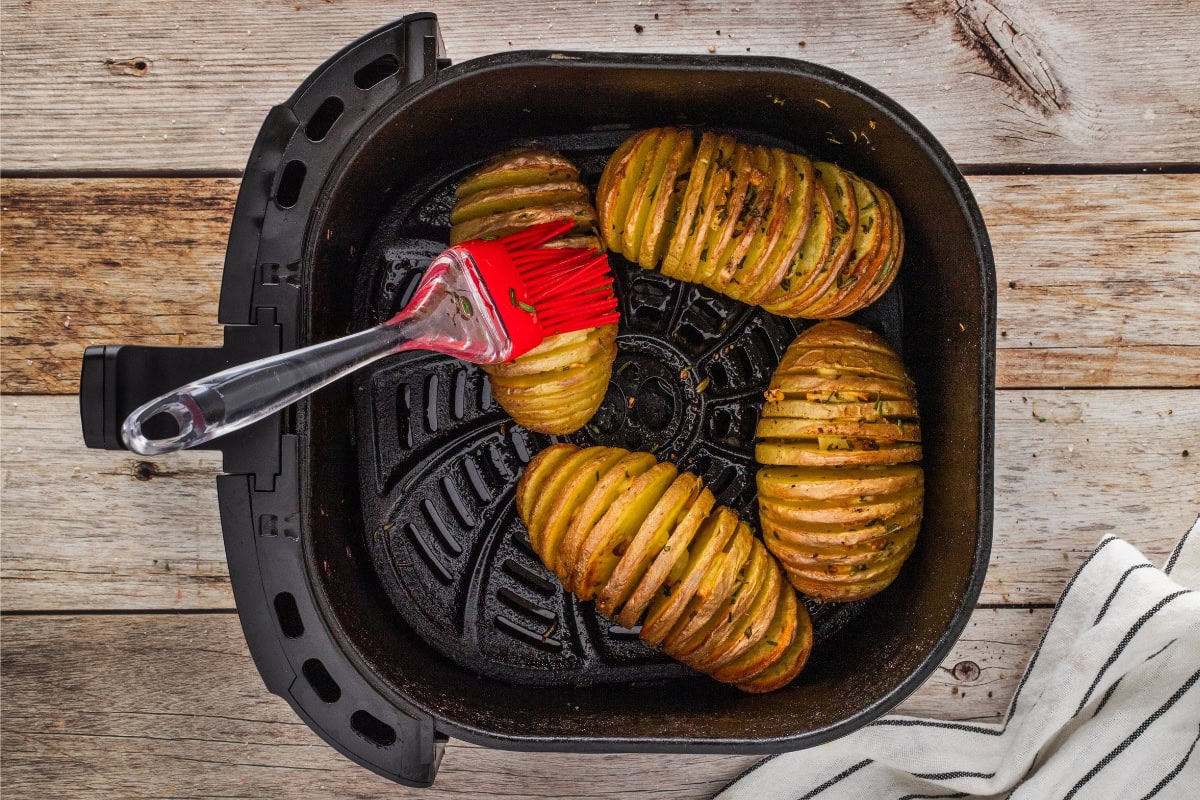 Hasselback potatoes and a brush inside an air fryer basket on a wooden table.