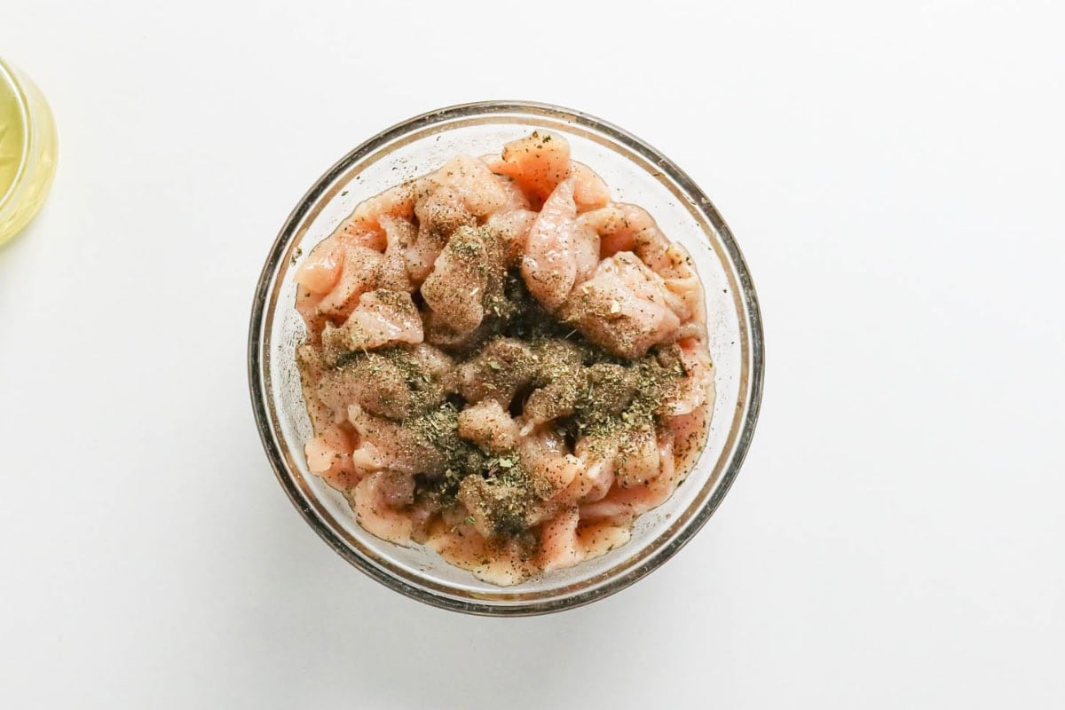 Glass bowl of raw, diced chicken with black pepper and spices on top.