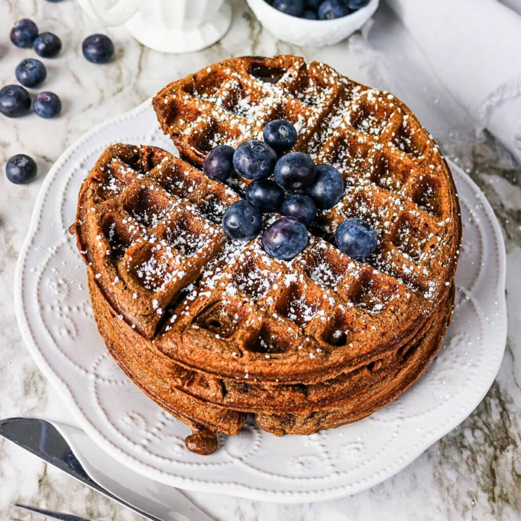 Stack of waffles topped with blueberries and powdered sugar on a white plate.