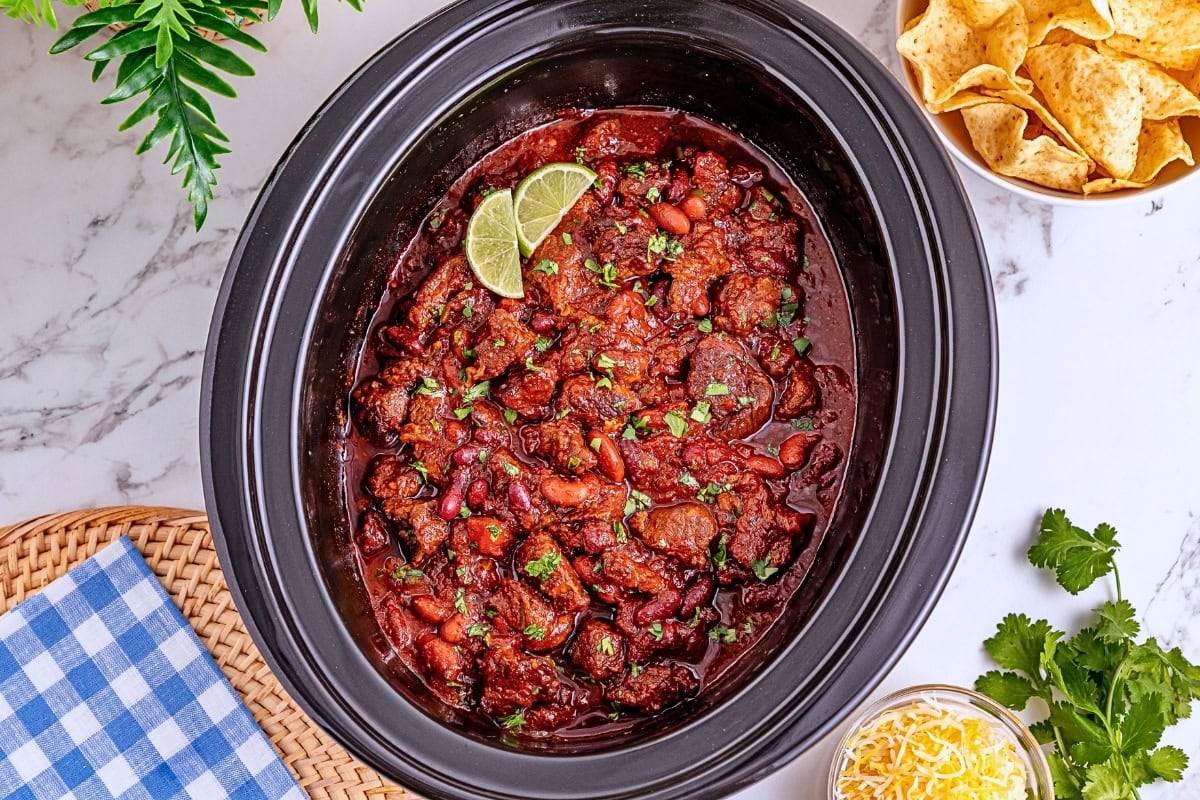 Slow cooker chili with beans and beef, garnished with lime, beside chips and cheese.