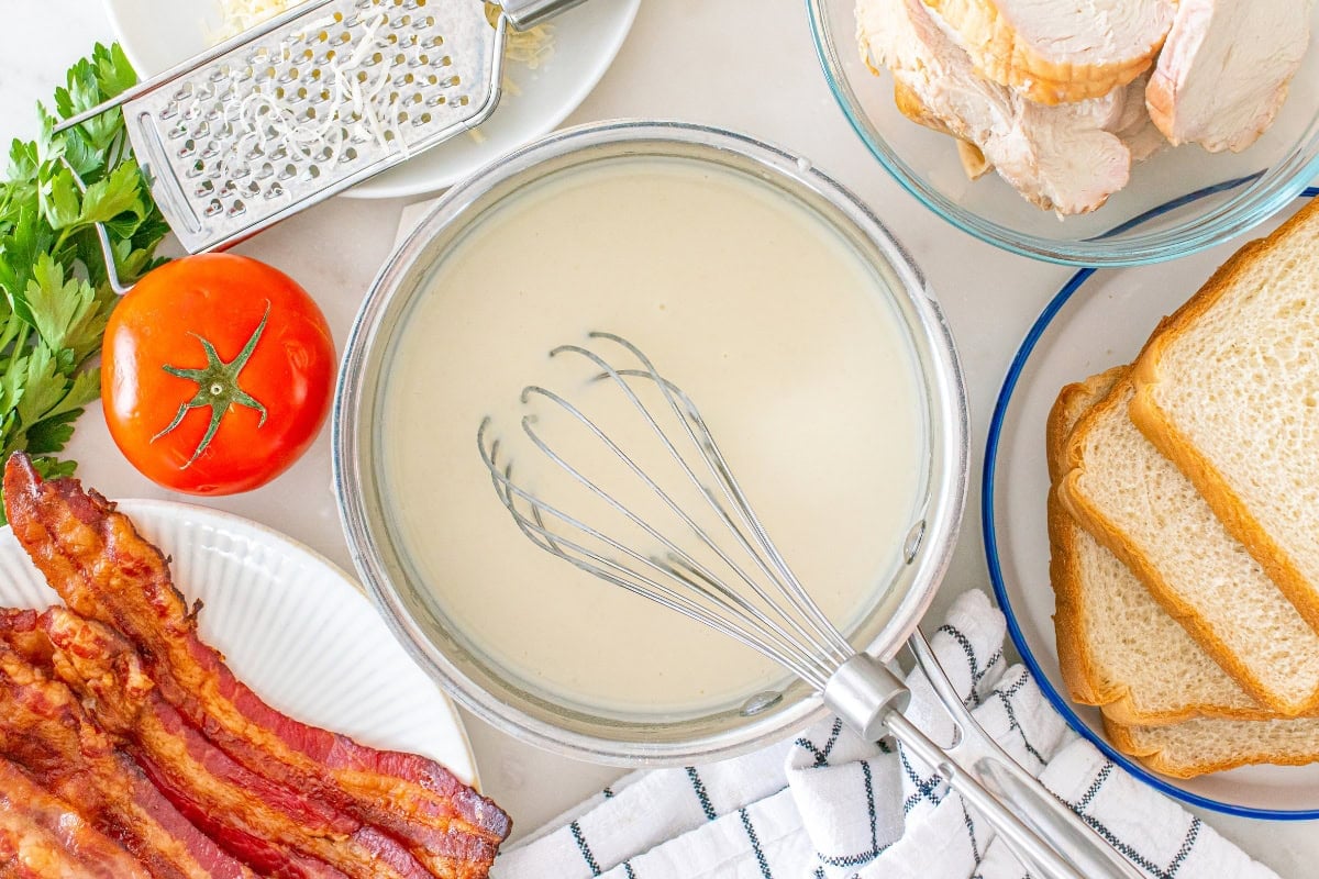 White sauce with whisk, surrounded by bacon, bread, turkey, tomato, and cheese.