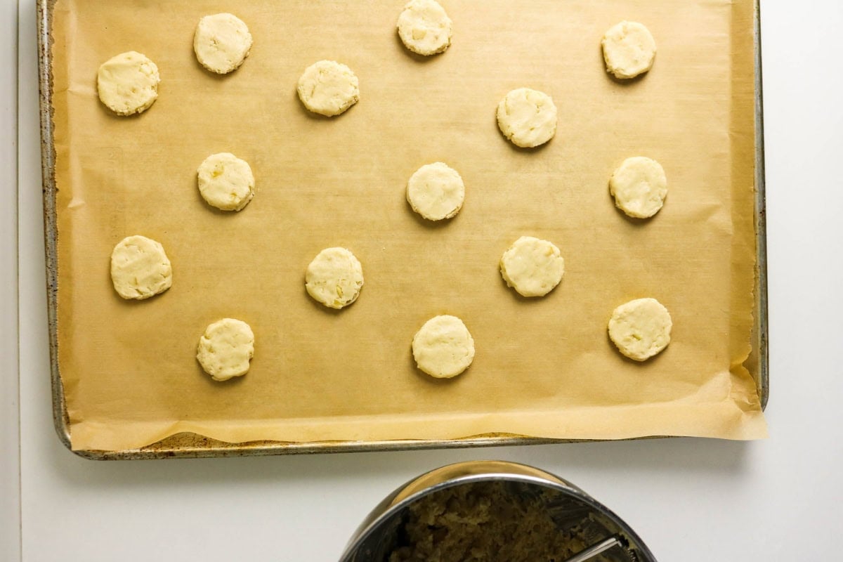 Raw cookie dough rounds spaced on parchment-lined baking sheet, bowl below.
