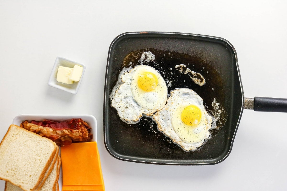 Sunny-side up eggs cook in a pan with bread, bacon, cheese, and butter nearby.