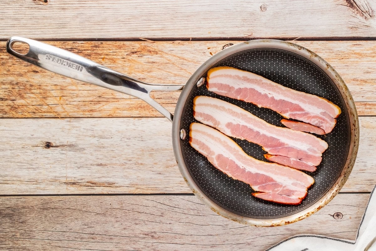 Three raw bacon strips in a frying pan on a wooden surface.