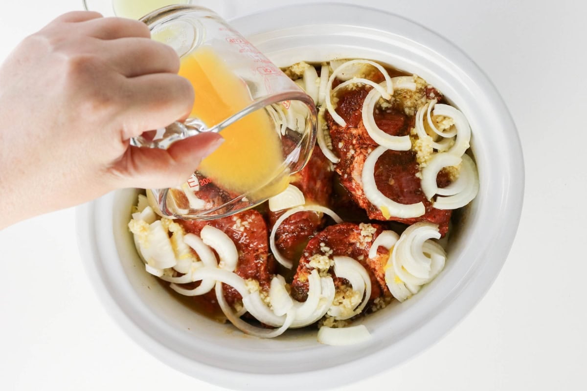 Hand pouring orange liquid into slow cooker with onions, sauce, and meat inside.
