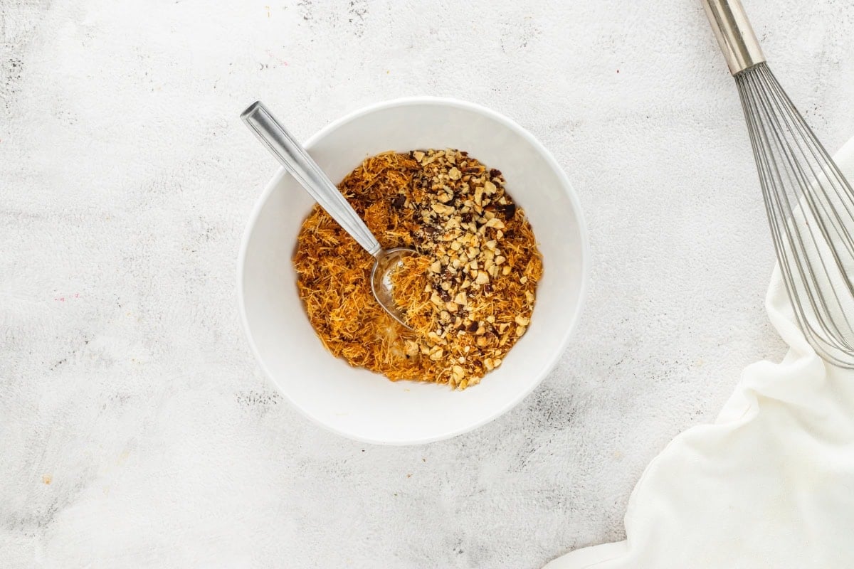 White bowl with shredded mixture and chopped nuts, spoon inside, whisk nearby.