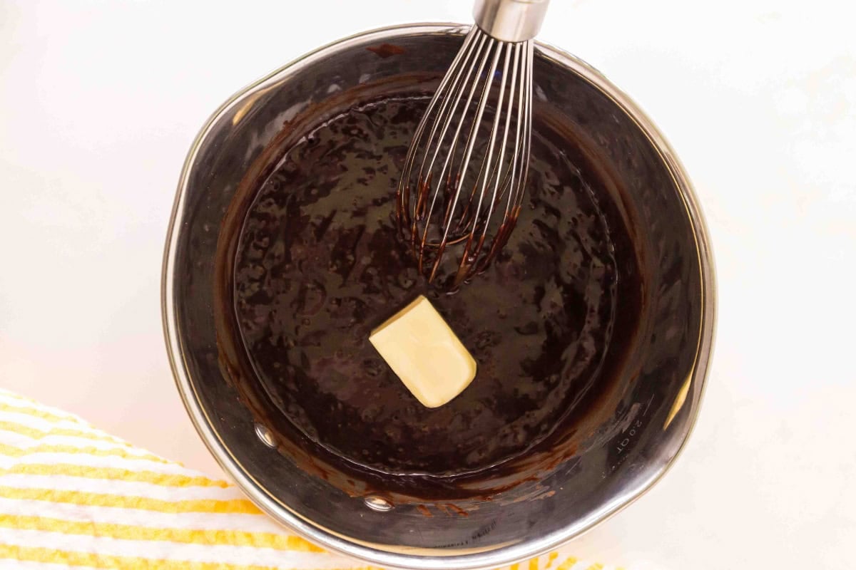 A whisk in a pot of chocolate mixture with a pat of butter melting in it.