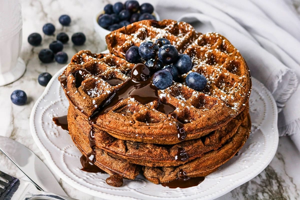 Three waffles stacked with blueberries, chocolate sauce, and powdered sugar.