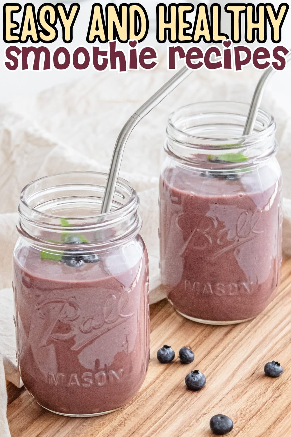 Two mason jars of purple smoothie with metal straws and blueberries on a wooden surface.