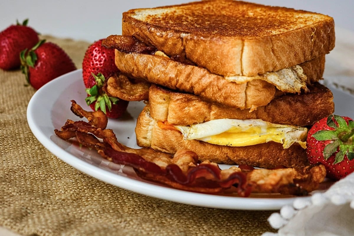 Stacked egg sandwiches with bacon and strawberries on a white plate.