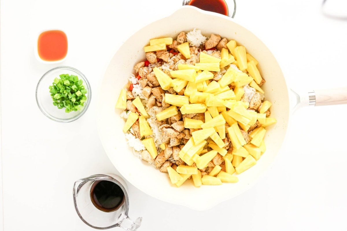 White pan with chopped pineapple, chicken, rice, and vegetables. Sauces nearby.