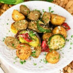 Roasted potatoes, sausage, zucchini, and peppers on a white plate with herbs.
