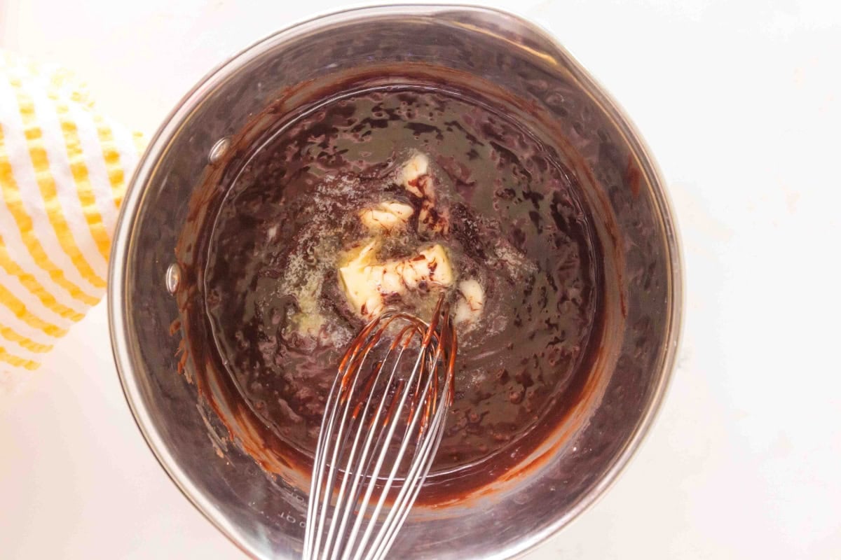 A whisk mixing butter into melted chocolate in a metal pot.