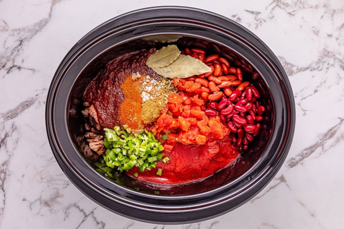 Slow cooker filled with beans, tomatoes, spices, bay leaf, and chopped peppers.