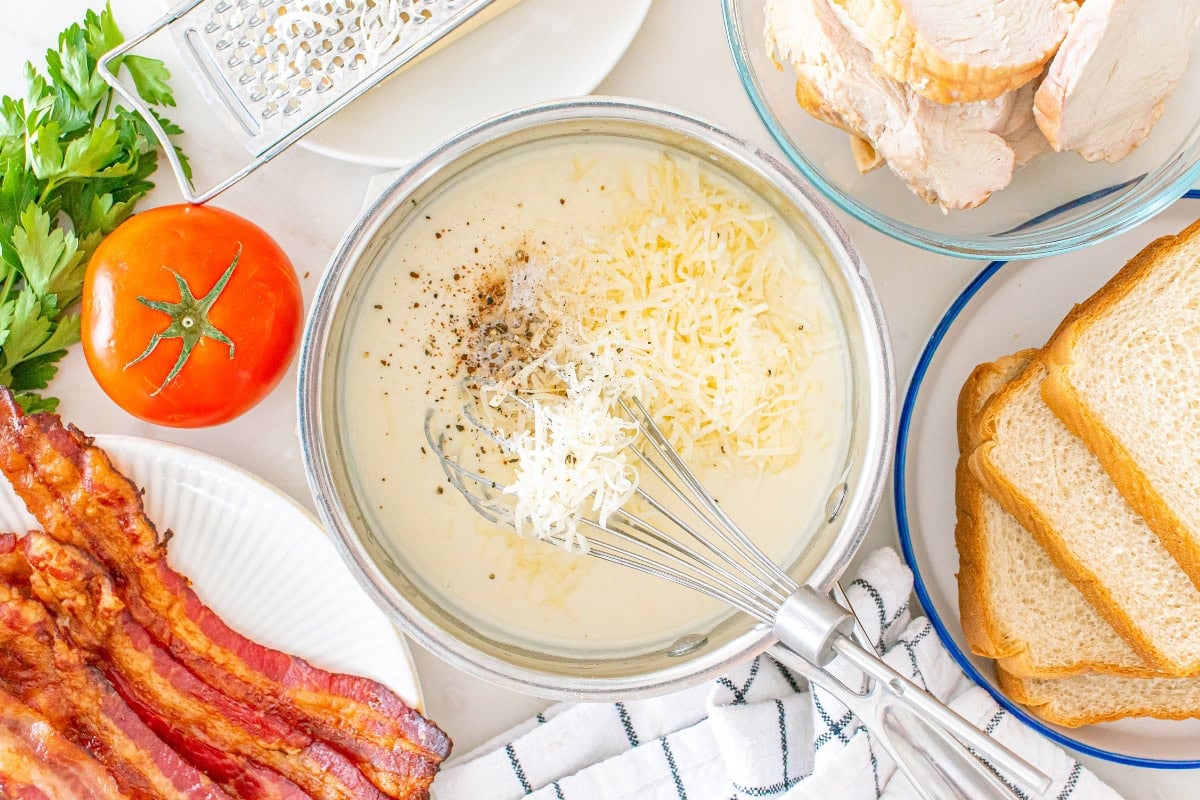 Whisking cheese into sauce, surrounded by bacon, bread, tomato, and chicken.