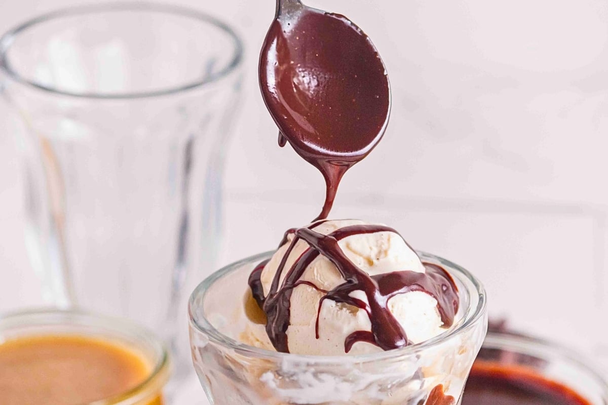 Spoon drizzling chocolate sauce over vanilla ice cream in a glass dish.
