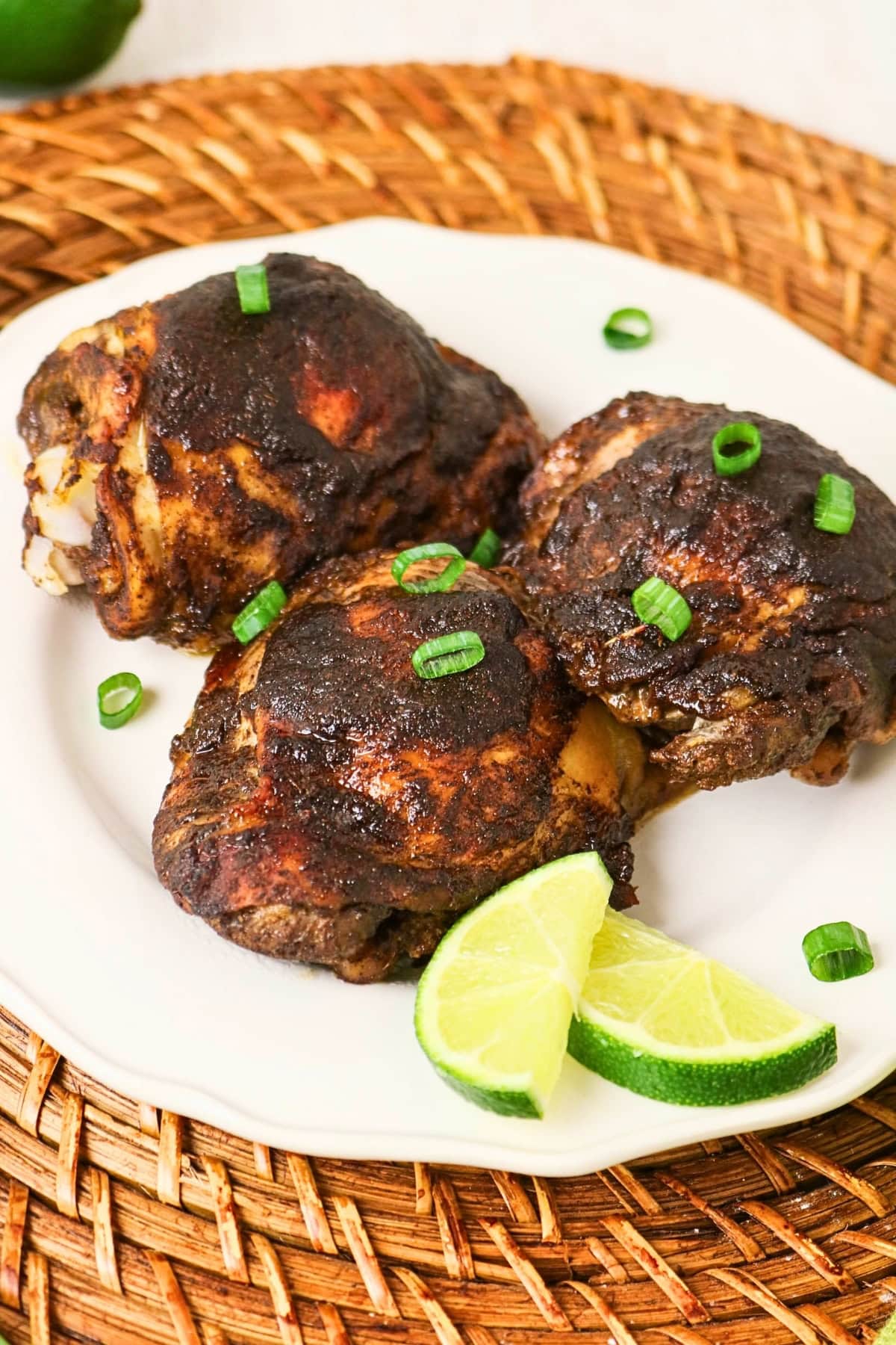 Three pieces of jerk chicken with lime wedges and green onions on a white plate.