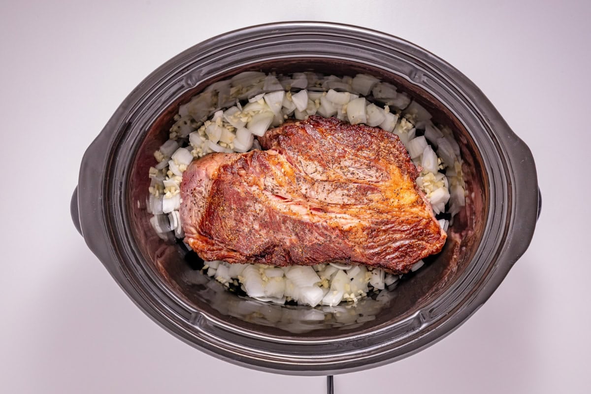 Browned beef roast on chopped onions in a slow cooker, ready to cook.