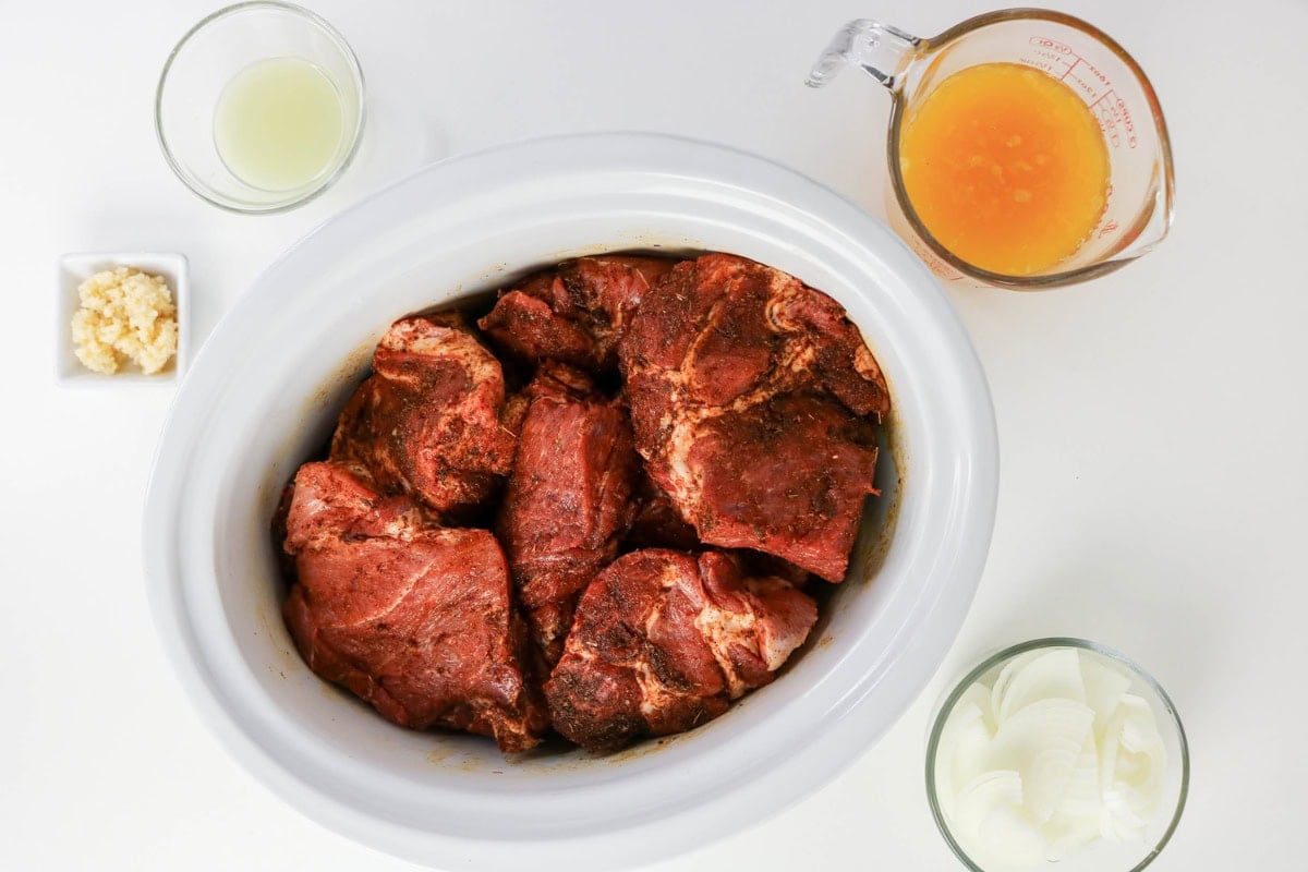 Seasoned pork in a slow cooker, with onion, garlic, juice, and broth nearby.