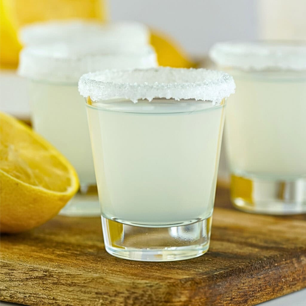 Three lemon drop shots with sugar rims on a wooden board, lemon halves nearby.