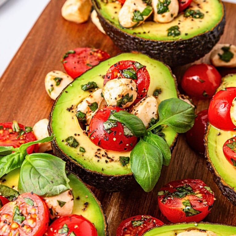 Halved avocados filled with tomatoes, mozzarella, herbs, and fresh basil on wood.