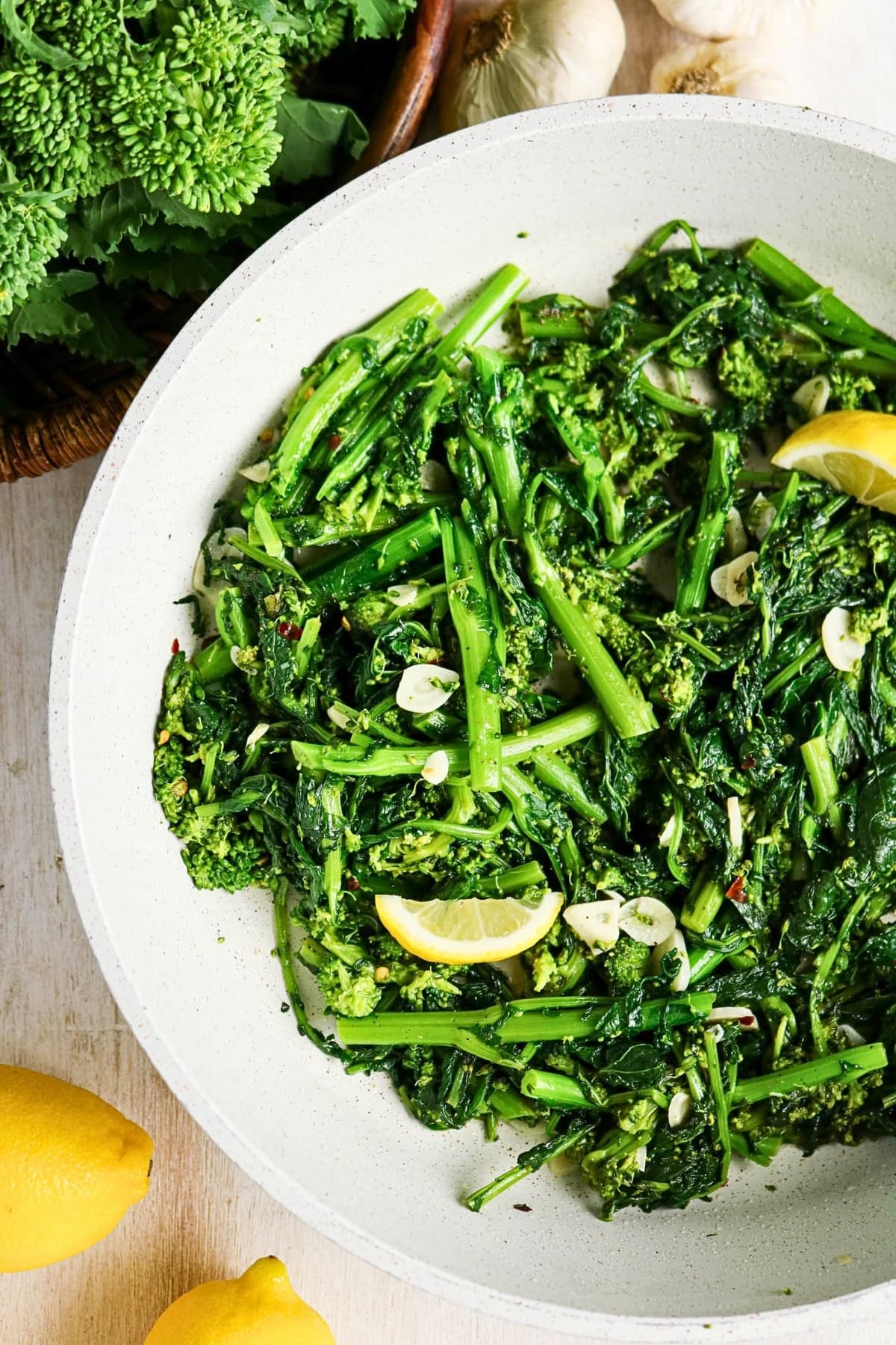 Cooked rapini with garlic and lemon wedges in a white pan.