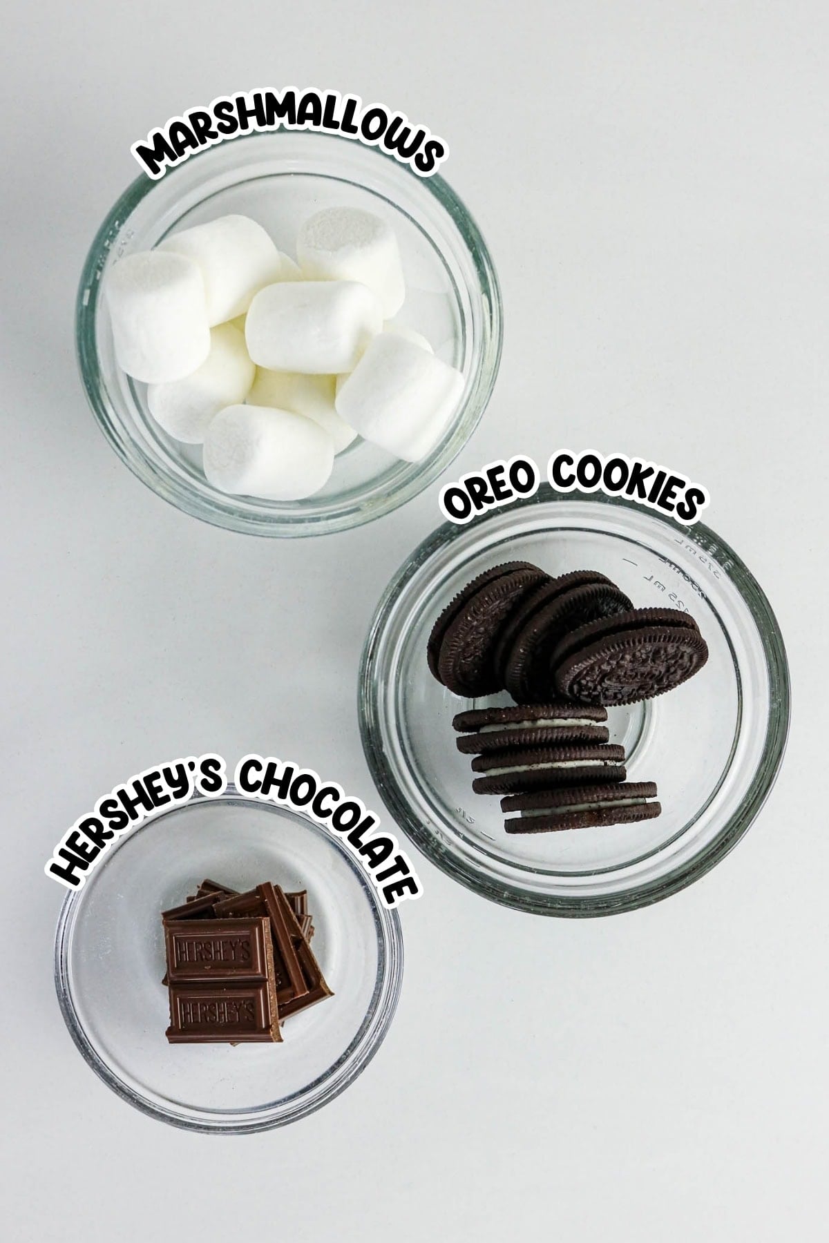 Bowls of marshmallows, Oreo cookies, and Hersheys chocolate on a white surface.
